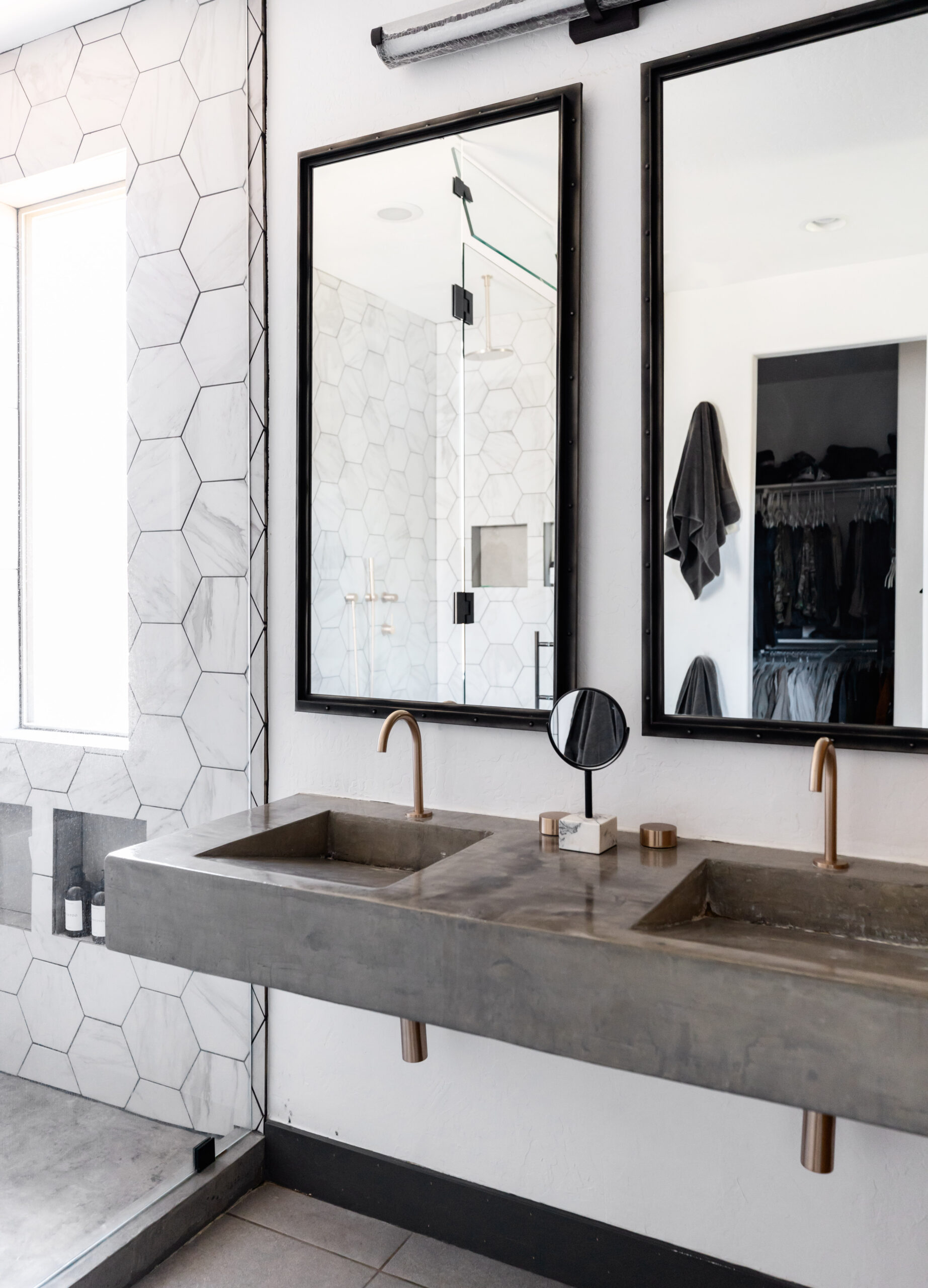 Accessible modern bathroom featuring a double floating concrete vanity with copper fixtures, dual mirrors, and exposed plumbing for wheelchair clearance. The space includes a walk-in shower with hexagonal tile and a sliding wood barn door, designed by Blue Copper Design.