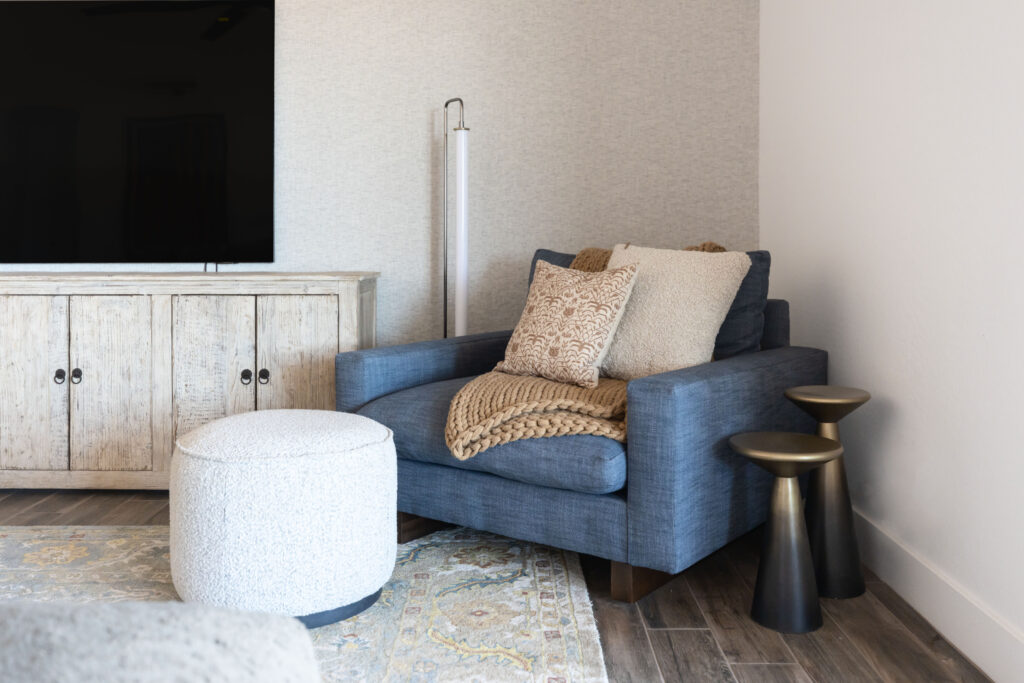 A cozy corner of a living room featuring a deep blue armchair with neutral throw pillows and a chunky knit blanket. Next to the chair are two modern metallic pedestal side tables. A white textured ottoman sits in front of the chair, and a rustic wood media console with a large flat-screen TV is positioned against the wall. The space is styled with warm tones and a soft patterned area rug on wood-look flooring.