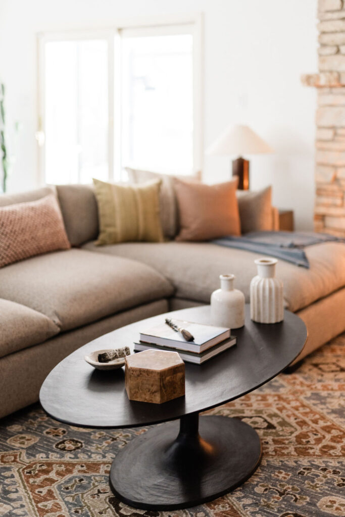 A modern living room featuring a taupe sectional sofa with textured throw pillows in muted tones. In front of the sofa is a black oval coffee table styled with ceramic vases, stacked books, and decorative objects. The room includes a patterned area rug, natural light from sliding glass doors, and a stone fireplace in the background.