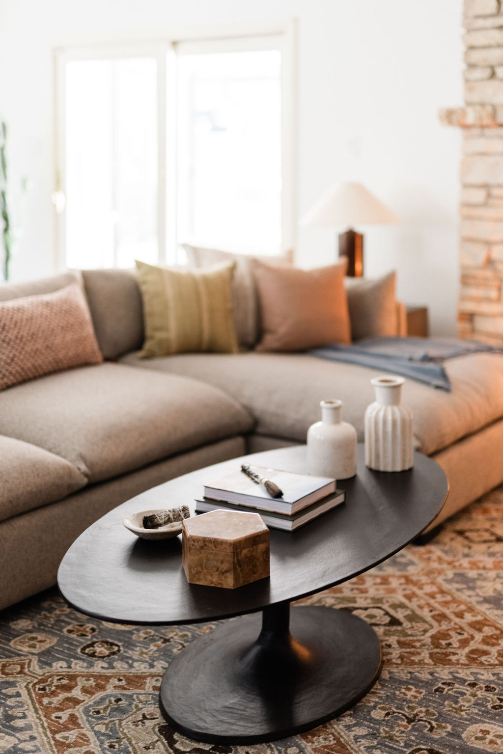 A modern living room featuring a taupe sectional sofa with textured throw pillows in muted tones. In front of the sofa is a black oval coffee table styled with ceramic vases, stacked books, and decorative objects. The room includes a patterned area rug, natural light from sliding glass doors, and a stone fireplace in the background.