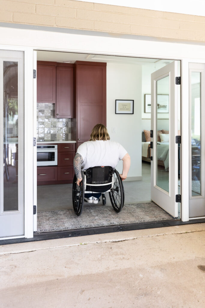 A wheelchair user enters a modern home through wide, step-free French doors that connect to an accessible kitchen with burgundy cabinets and open floor space.