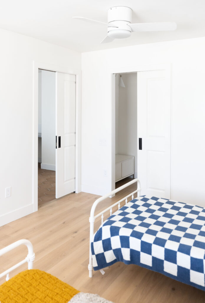Bright and airy bedroom featuring two white sliding pocket doors, white walls, light wood flooring, and a bed with a bold blue and white checkerboard blanket.