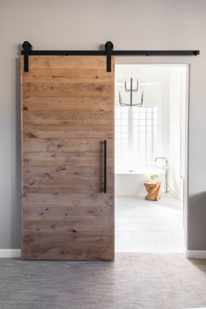 Modern wood barn door with black hardware partially open to reveal a bright, accessible bathroom with white tile flooring, a freestanding tub, and a sculptural wood side table.