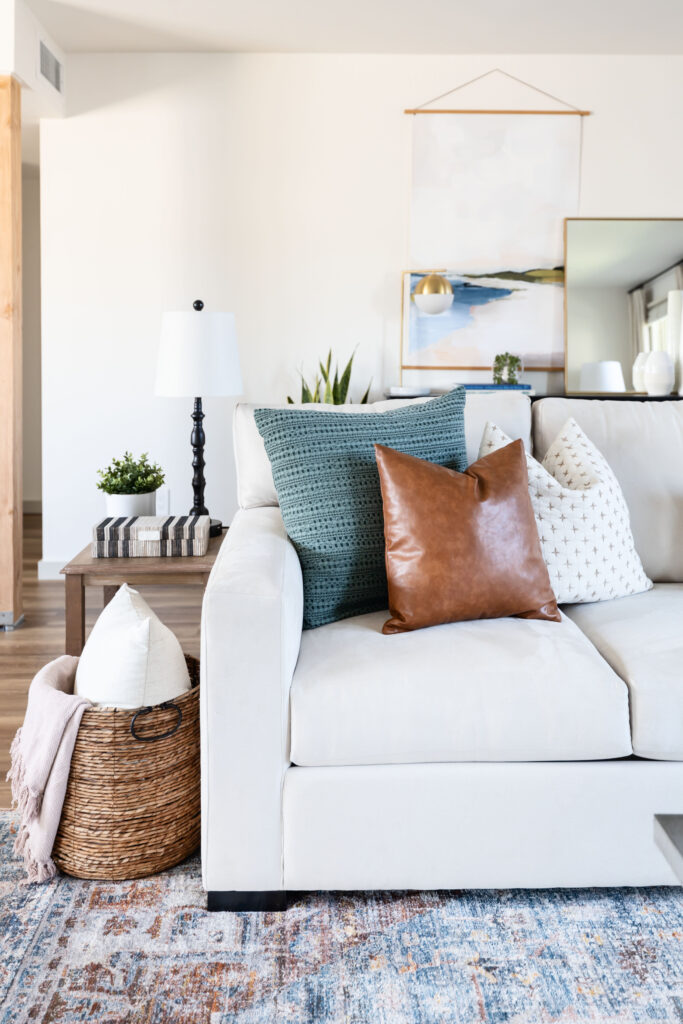 Cozy living room corner in an accessible apartment featuring a neutral sectional sofa with colorful throw pillows, a woven basket with a blanket and pillow, a wood side table with a black lamp, and layered artwork and greenery for a warm, inviting look.