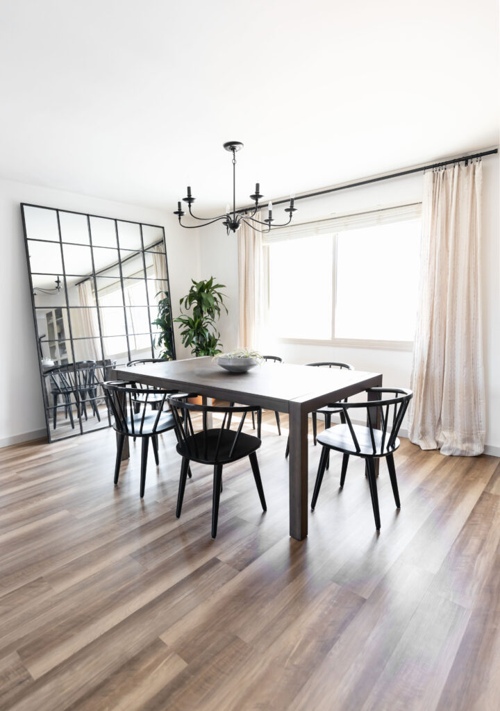 Modern dining area inside an accessible apartment featuring a black dining table with matching chairs, wood-look flooring, a large grid mirror, and soft natural light from a wide window with floor-length curtains.