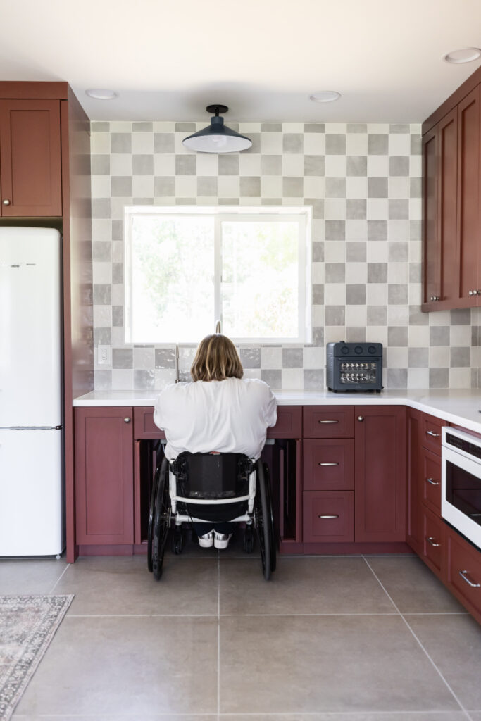 Person using a wheelchair positioned at a roll-under kitchen sink with red cabinetry, checkerboard tile backsplash, and a window above the sink.