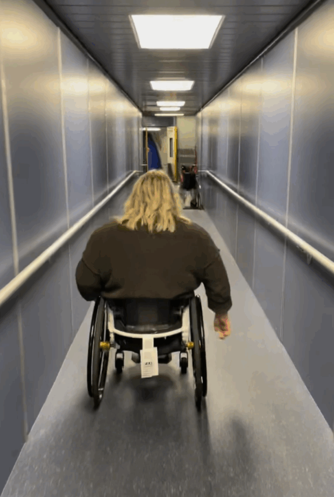 A traveler in a wheelchair with blonde hair moves independently down a narrow airport jet bridge toward the airplane, representing accessible air travel tips.
