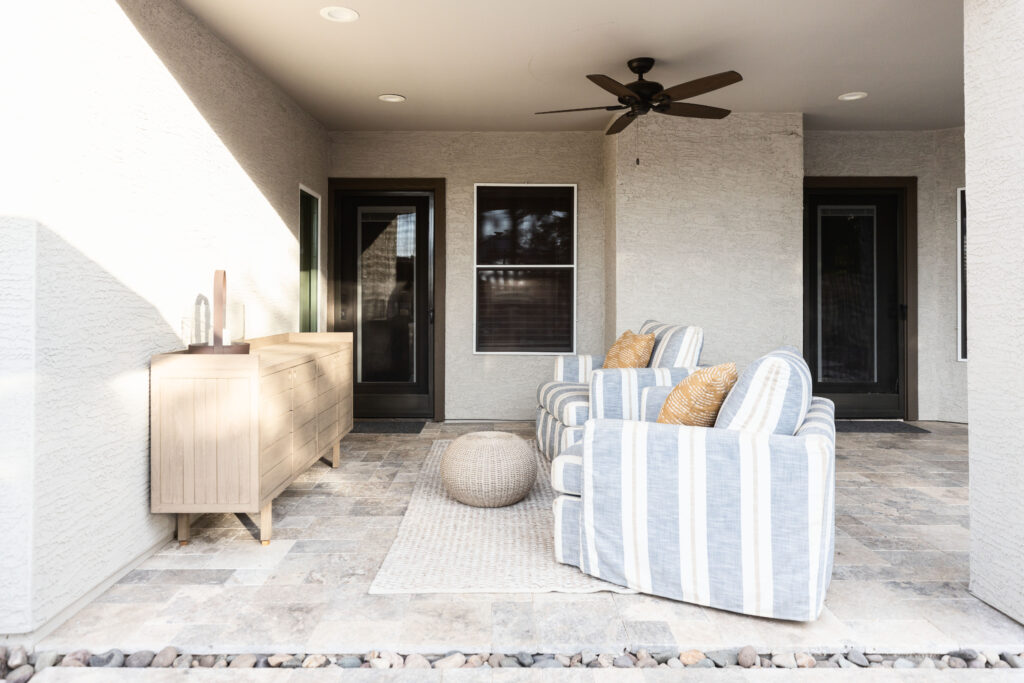 Covered patio with light stone tile flooring, two striped upholstered chairs with gold accent pillows, a woven ottoman, and a light wood sideboard. Ceiling fan and natural sunlight enhance the relaxed, barrier-free outdoor setting. Designed by Blue Copper Design.
