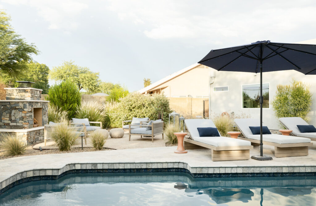 Backyard pool area with cushioned lounge chairs, navy pillows, and a large black umbrella on a stone deck. Features desert landscaping with native plants, a stone outdoor fireplace, and comfortable seating areas. Designed by Blue Copper Design for inclusive outdoor living.