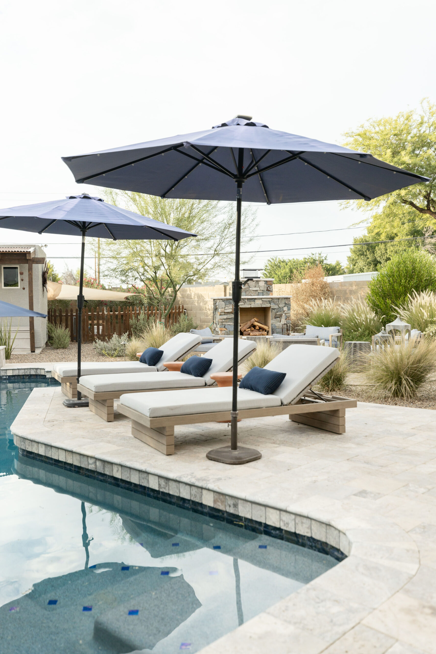 Poolside area featuring cushioned lounge chairs with navy pillows under large blue umbrellas, set on a light stone paver deck. Reflections in the water enhance the serene, accessible outdoor design. Surrounded by desert landscaping and a stone outdoor fireplace in the background. Designed by Blue Copper Design.
