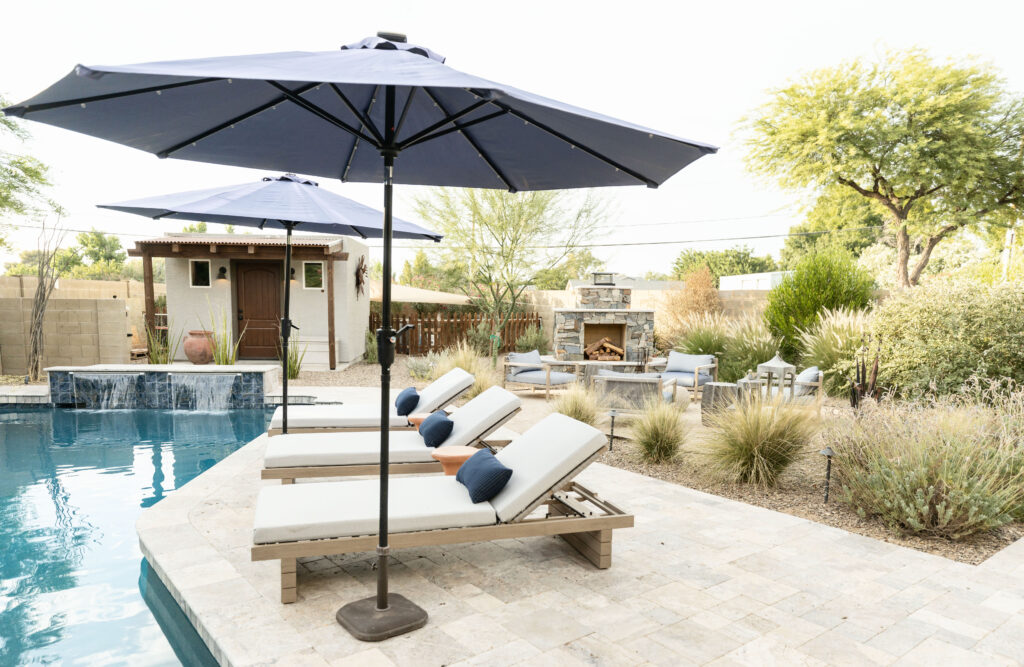 Accessible poolside area with cushioned lounge chairs, navy accent pillows, and a large black umbrella on a stone paver deck. Background features a rustic stone fireplace, native desert landscaping, and a small stucco pool house. Designed by Blue Copper Design.