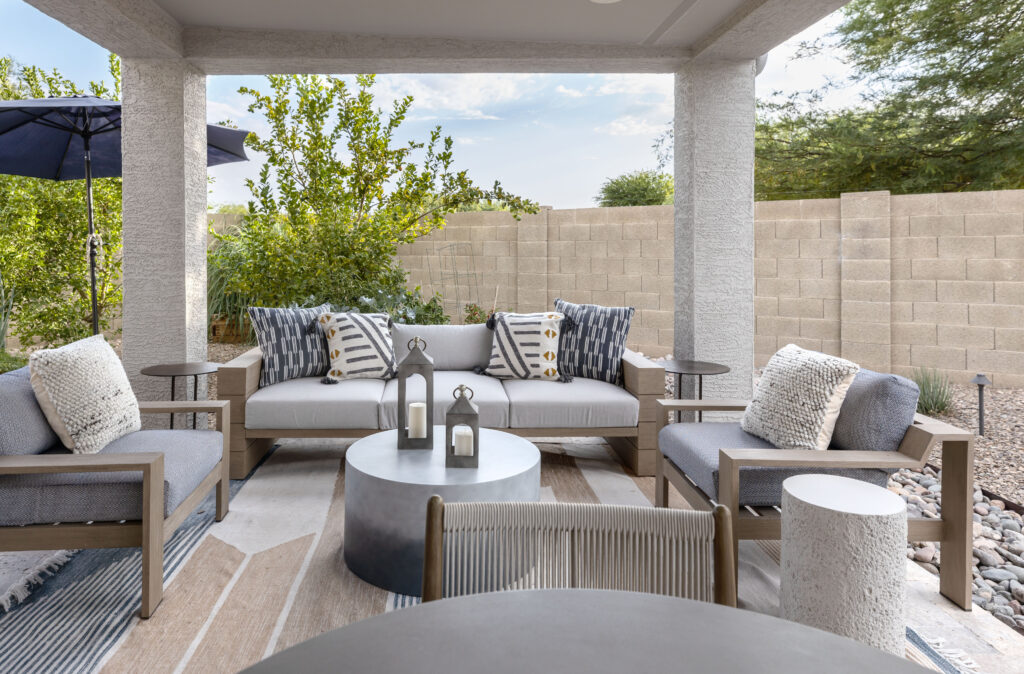 Modern covered patio featuring accessible seating with neutral-toned cushioned chairs and sofa, decorative throw pillows, round coffee table with candle lanterns, and a textured area rug. Surrounded by desert landscaping, block wall, and shaded by a navy umbrella. Designed by Blue Copper Design.