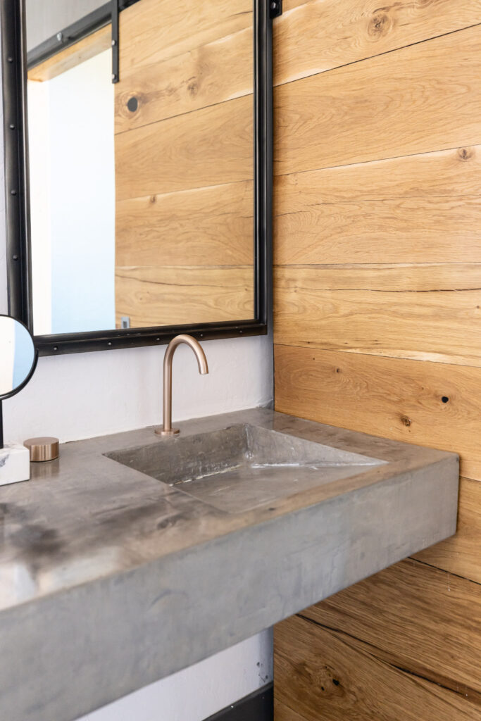 Modern bathroom vanity with a concrete sink, sleek brushed metal faucet, black-framed mirror, and warm wood plank wall for a rustic yet contemporary look.
