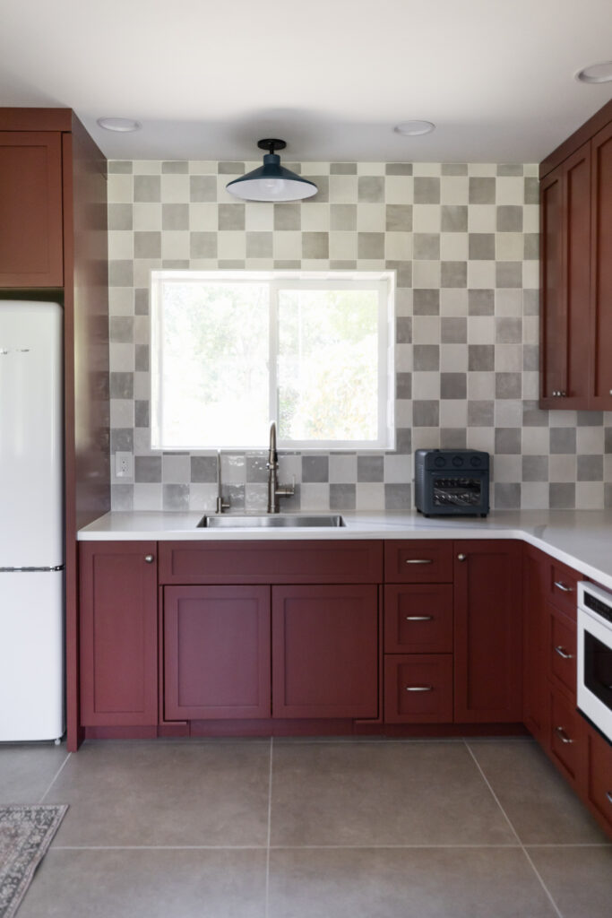 Modern kitchen with deep red cabinetry, stainless steel sink, gray checkered tile backsplash, and white countertops, accented by a black-and-white pendant light.