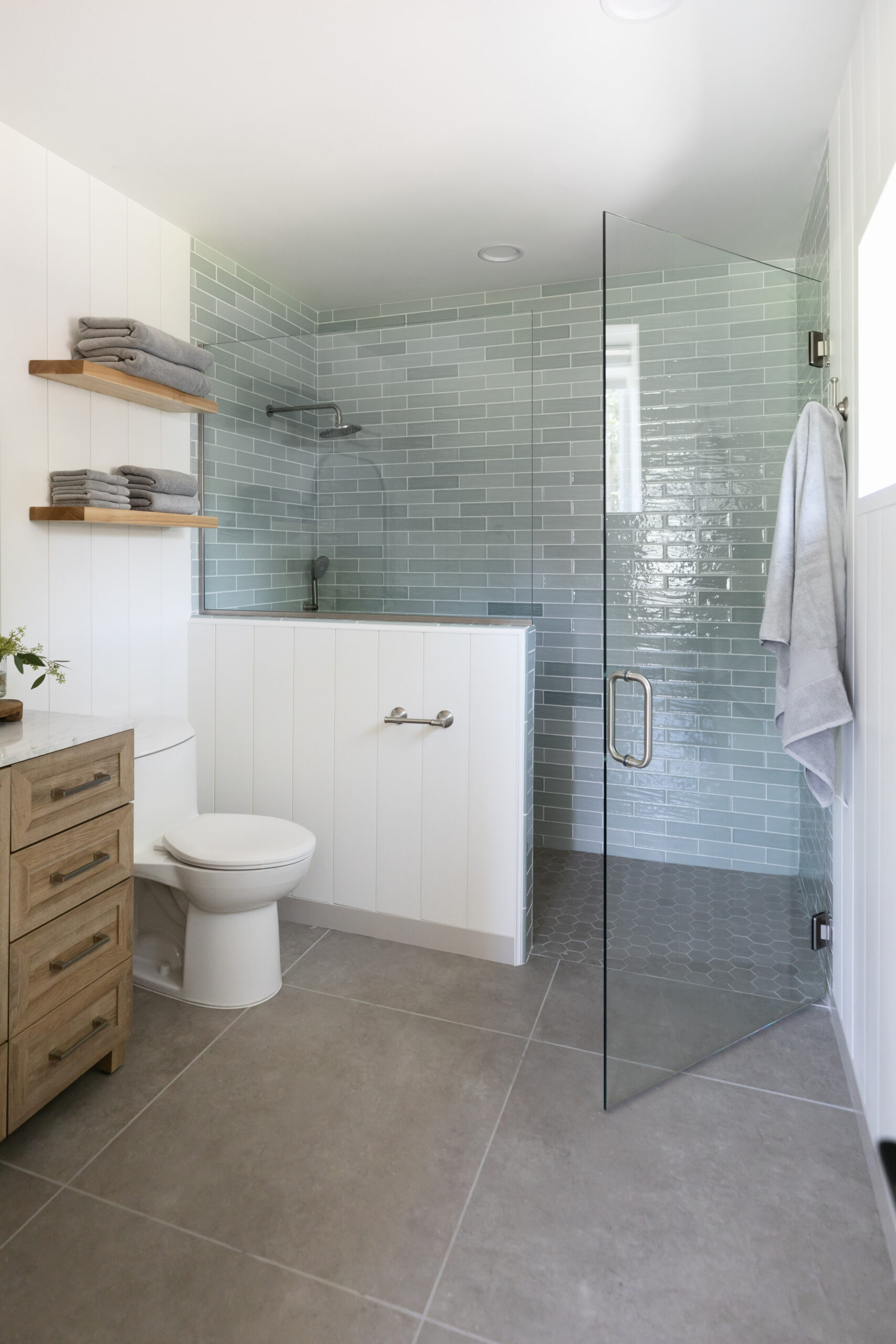 Modern accessible bathroom with a roll-in shower featuring glass doors, light green subway tile, and nearby grab bar next to the toilet.