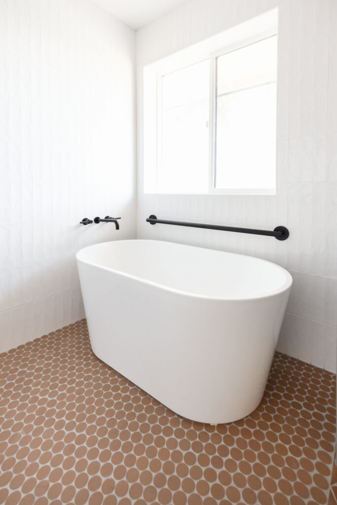 Freestanding white bathtub with matte black grab bar on a white tiled wall, showcasing accessible and stylish bathroom design by Blue Copper Design.