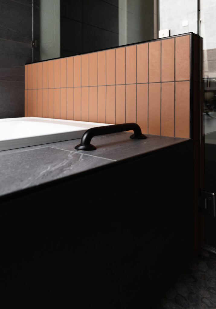 Close up of grab bar on built in bathtub with matte black grab bar on a orange tiled wall, showcasing accessible and stylish bathroom design by Blue Copper Design.