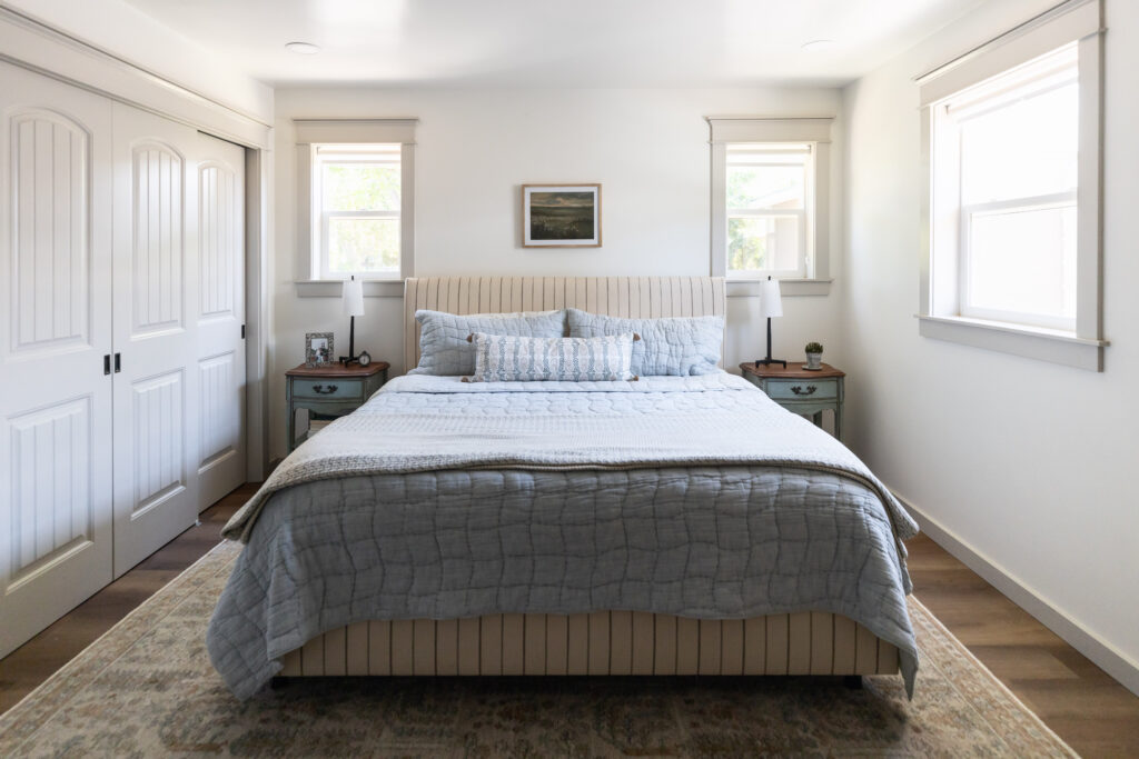 Primary bedroom with king bed centered between two windows. The bed is upholstered in cream striped fabric and has a blue quilt on the bed. 