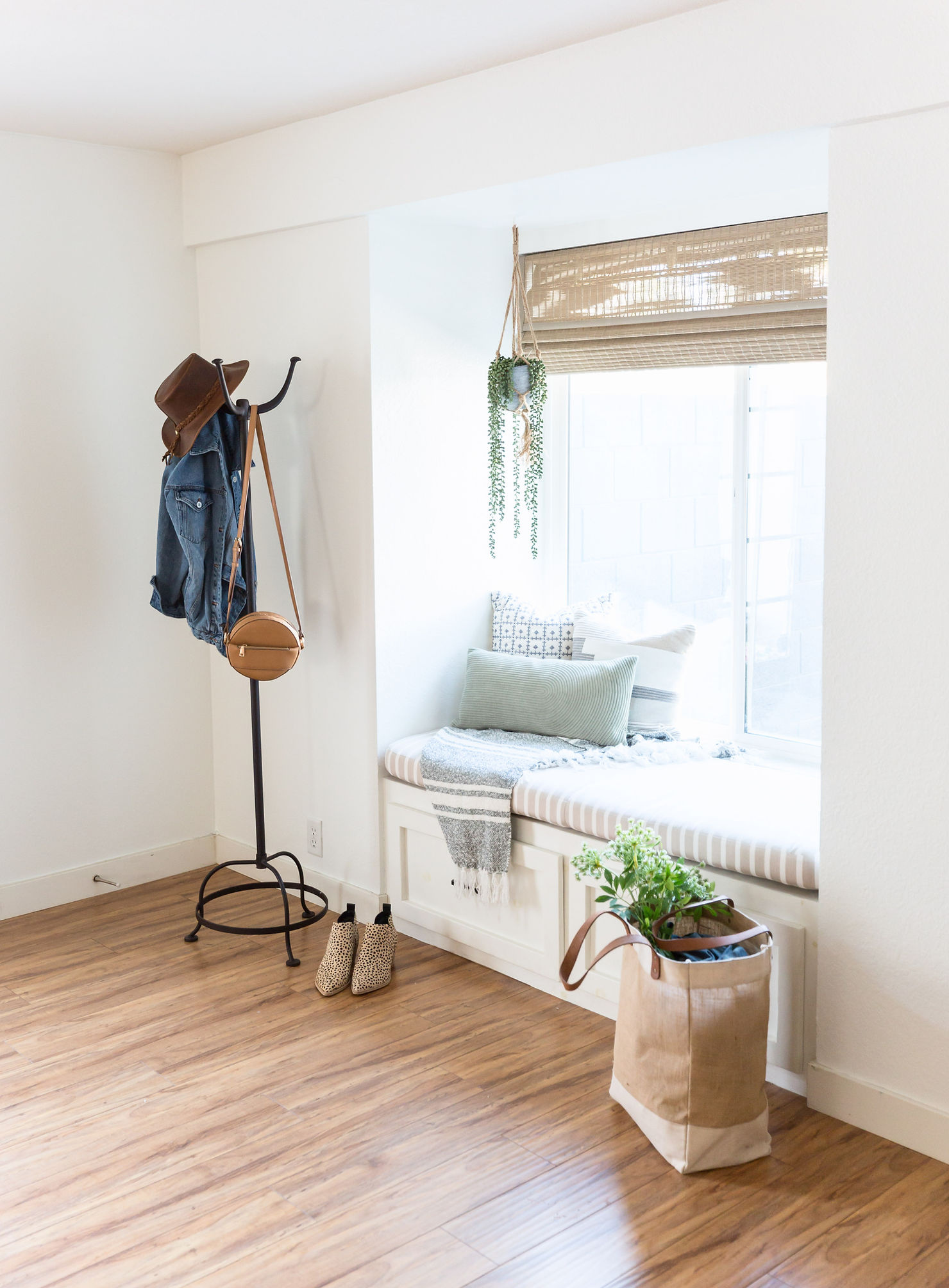 Bright and cozy entryway with a built-in window seat, layered throw blanket, and neutral pillows. A woven hanging plant and bamboo shade add natural texture, while a coat rack with a hat and denim jacket, patterned boots, and a canvas tote with greenery complete the inviting “homey” design.