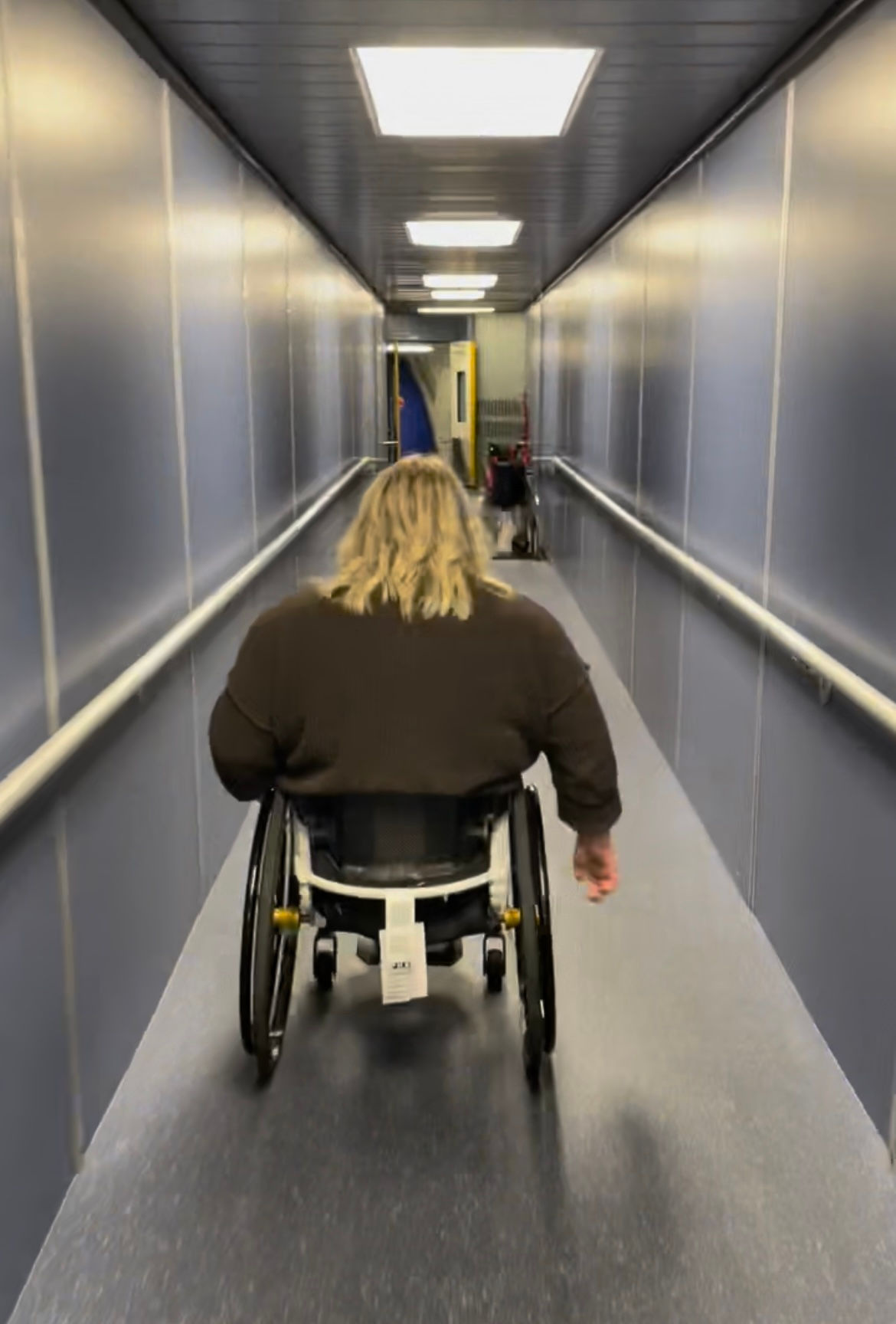 A traveler in a wheelchair with blonde hair moves independently down a narrow airport jet bridge toward the airplane, representing accessible air travel tips.