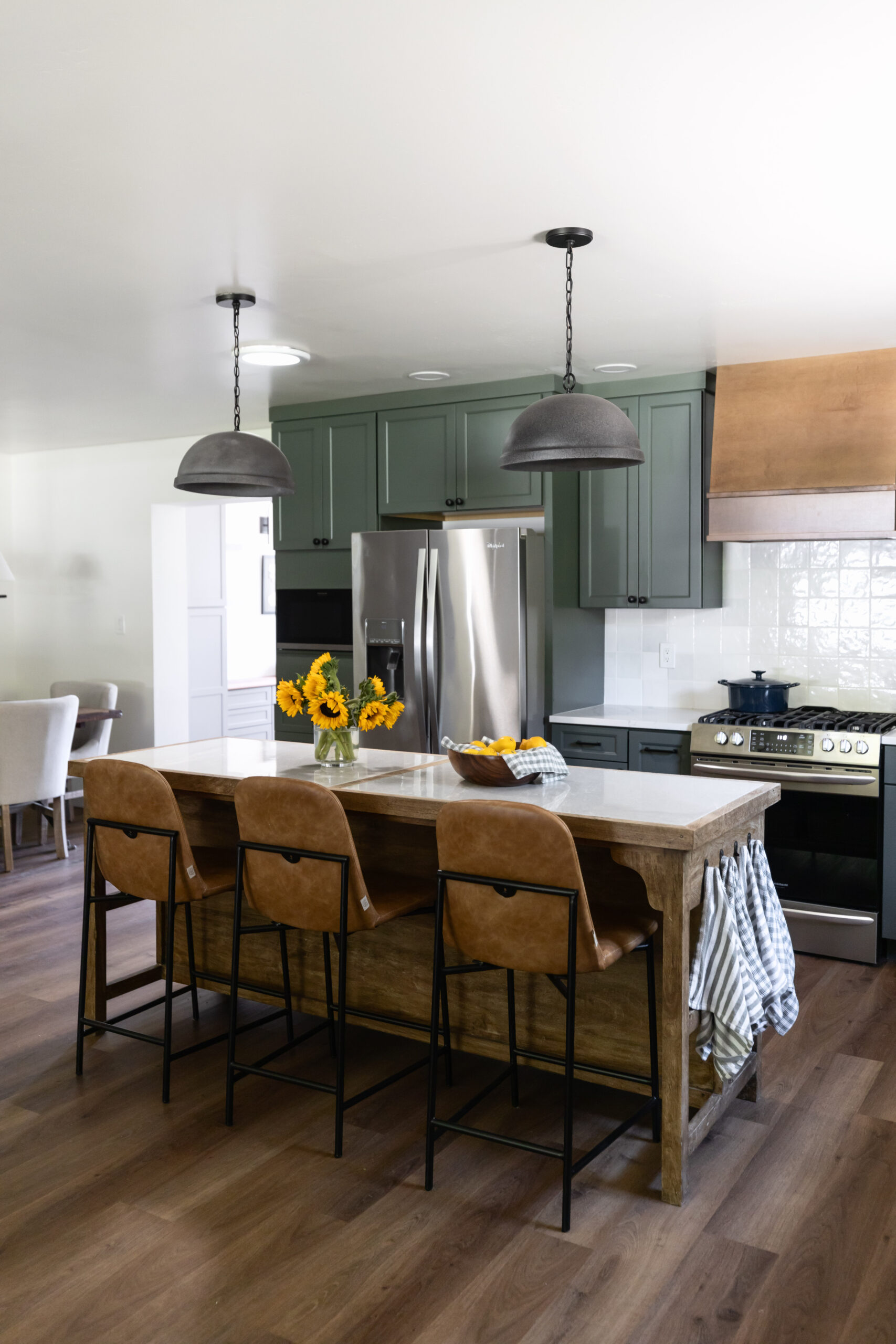Green kitchen cabinets with wood island with three leather barstools and two concrete pendants