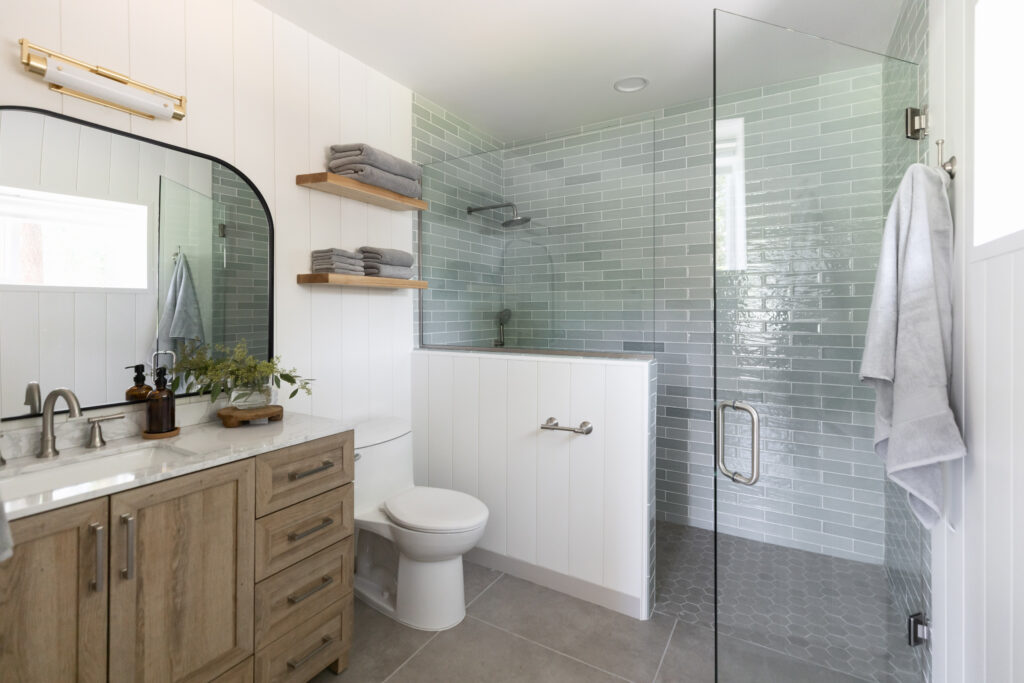 Barrier free bathroom with curb less shower, blue subway tile, wood vanity with white marble countertop, black mirror above the vanity, gold wall sconce.  