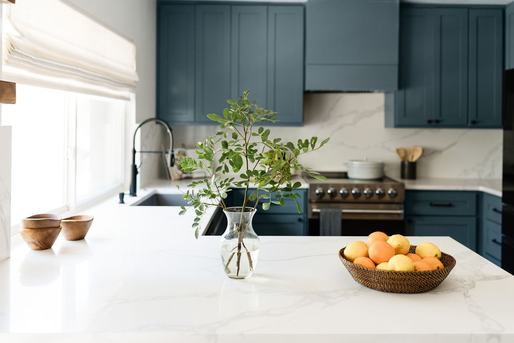 Close up kitchen with white quartz countertop, blue cabinets, stainless steel stove and oranges in a woven bowl. Achieved with a designer and contractor