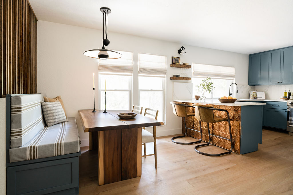 Open concept kitchen and dining area achieved with a designer and contractor. Kitchen has blue cabinets, white countertop, copper tiles, and leather counter stools. Dining room has built in bench and wood table. 