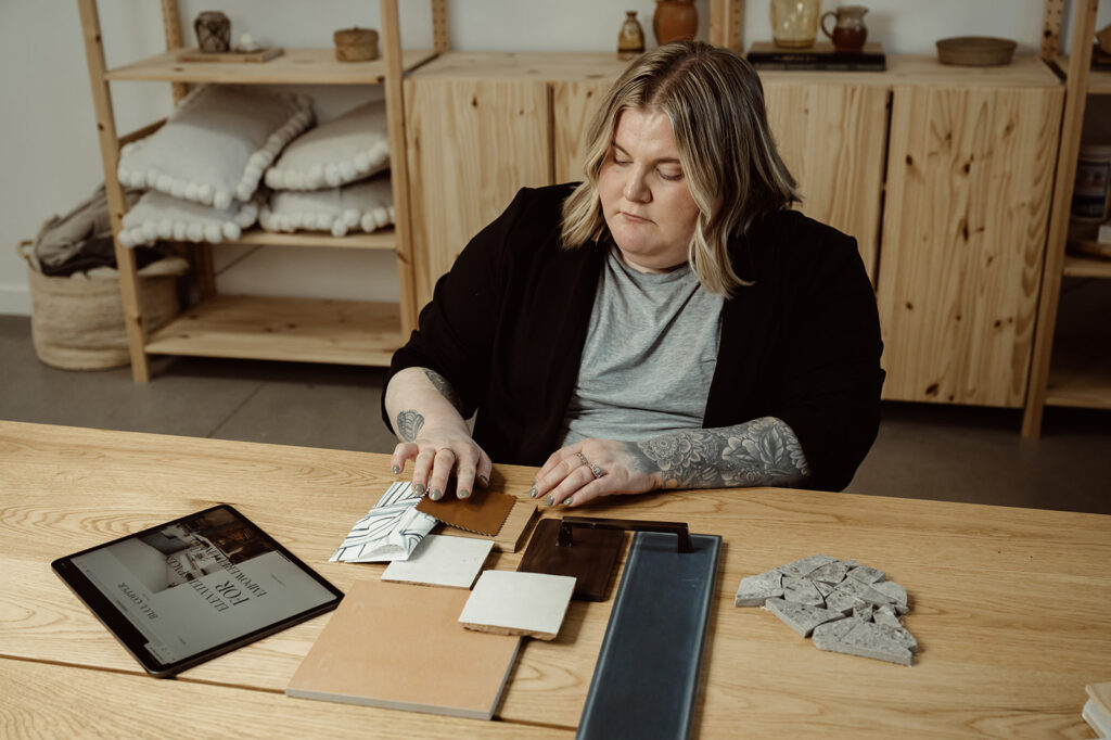 Maegan wearing a grey shirt, black blazer with short blonde hair looking at fabrics and materials on a wood desk with an iPad on the desk.