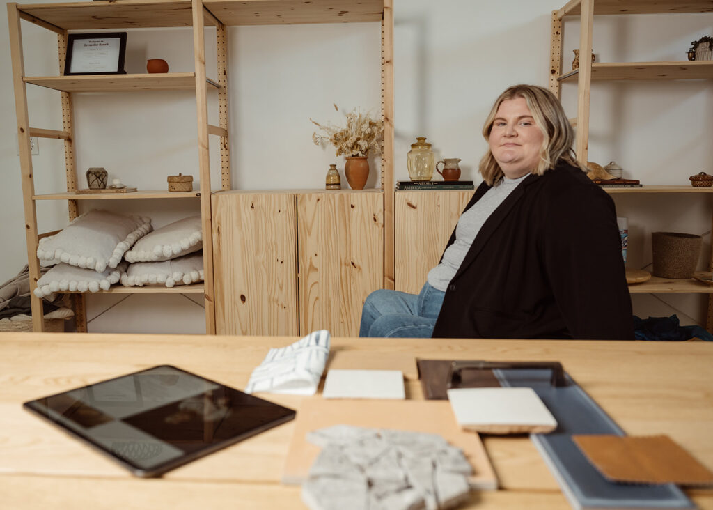 Maegan is an interior designer has short blonde hair, wearing a black blazer and blue jeans behind a wood table with fabric and material samples on the table.
