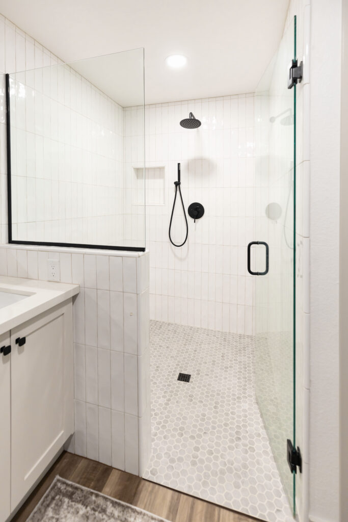 Accessible roll-in shower featuring a zero-threshold entry, white vertical subway tile walls, slip-resistant hexagon floor tiles, a black handheld shower head, and frameless glass door.