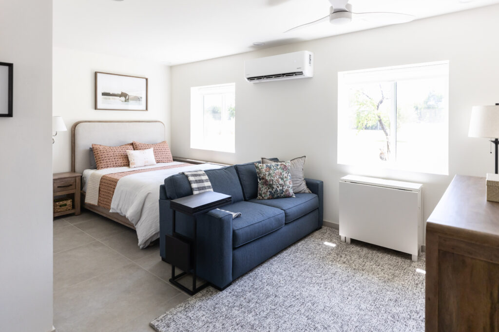 Accessible Barrier Free casita bedroom with blue pull out sofa, grey rug, wood queen size bed, white walls, and large windows. 