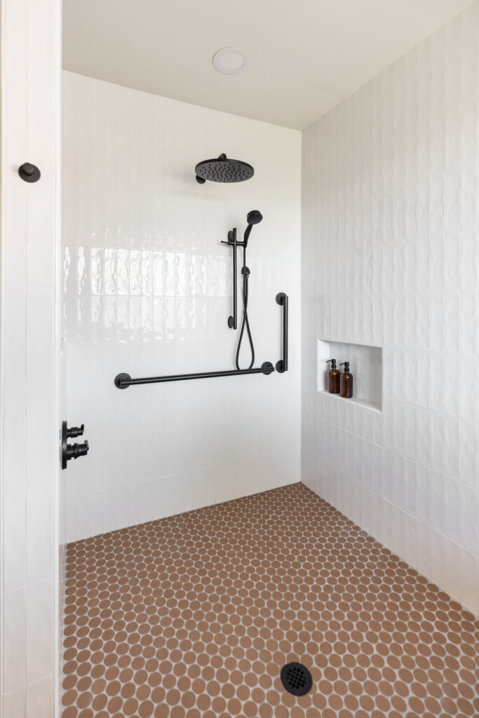 Accessible roll-in shower with white vertical tile walls, matte black grab bars, a handheld shower head on a slide bar, a rainfall shower head, brown penny tile flooring, and a built-in wall niche with amber soap dispensers.