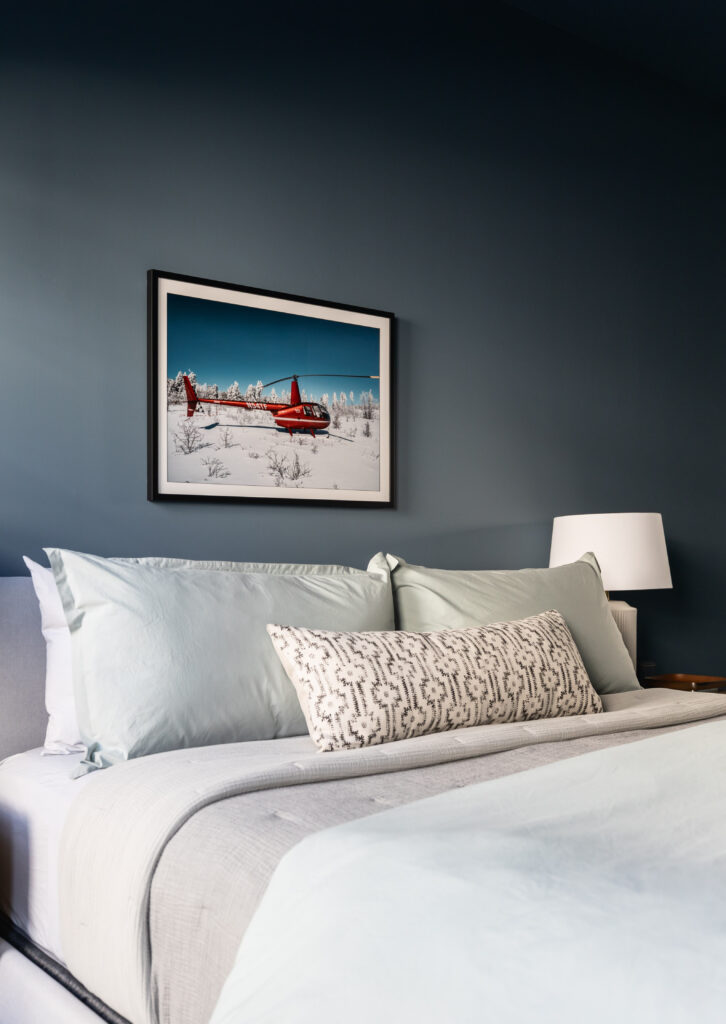 Moody bedroom with dark blue paint, light grey bedding, with a geometric patterned pillow on the bed. Above the bed is a picture of a red helicopter in the white snow with a blue sky. 