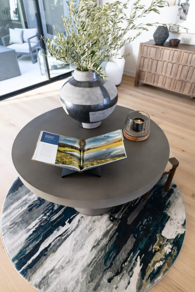 Grey concrete table in bright room with round rug on light hardwood floors. Concrete table is styled with an open book, large plant in ceramic vase and candle. 
