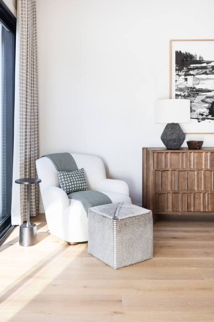 Bright and modern reading nook featuring a plush white armchair with a green throw and patterned pillow, positioned beside a large window with geometric curtains. A soft cube ottoman and black metal side table complete the space, with a warm wood credenza and abstract black and white artwork in the background.