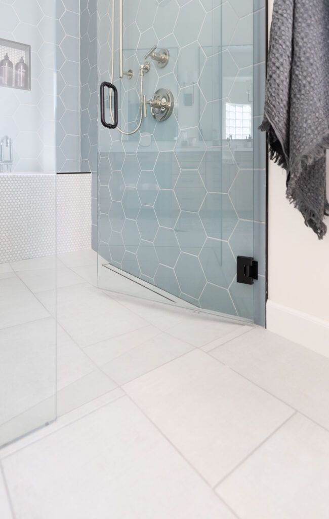 Accessible shower with large white floor tiles, blue hexagon wall tiles, linear drain at the zero-threshold entry, brushed nickel shower fixtures, and a frameless glass door.