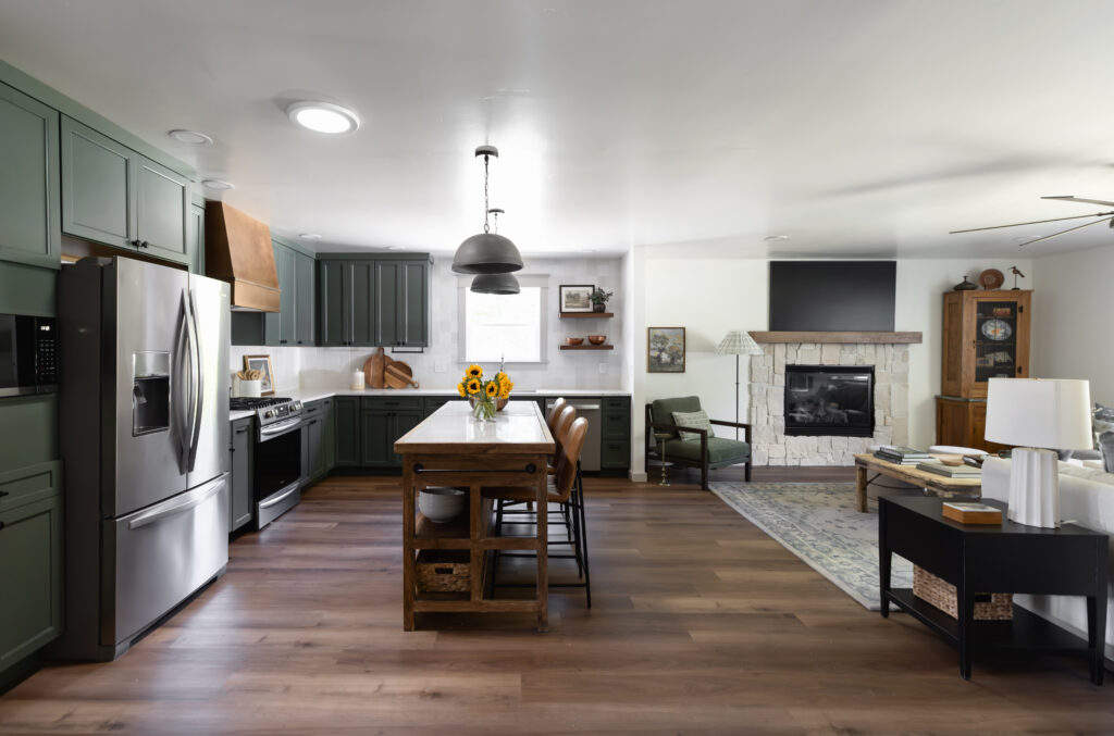 Open-concept kitchen and living area featuring sage green cabinetry, stainless steel appliances, and a rustic wood island with seating. The kitchen flows into a cozy living room with a stone fireplace, mid-century style furniture, and a mix of warm wood tones and soft textiles.