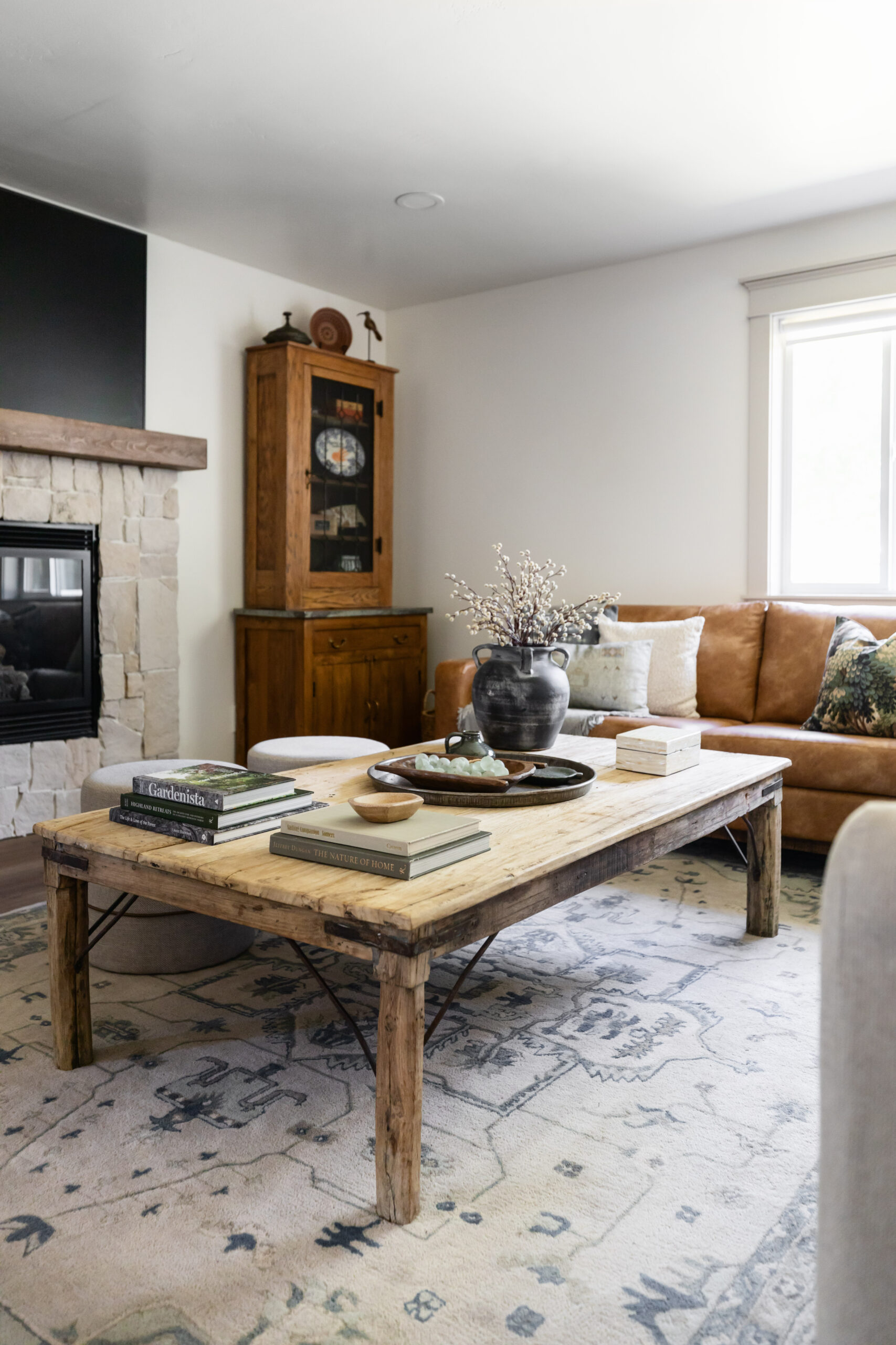 Cozy living room corner featuring a rustic wood coffee table styled with books and decor, a caramel leather sofa with layered pillows, a vintage wood cabinet, and a stone fireplace with a wood mantel.