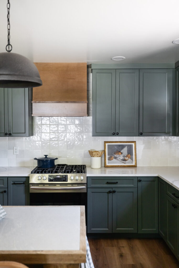 Green kitchen cabinets with white tile backsplash, wood range hood, concrete pendant, gas range stove