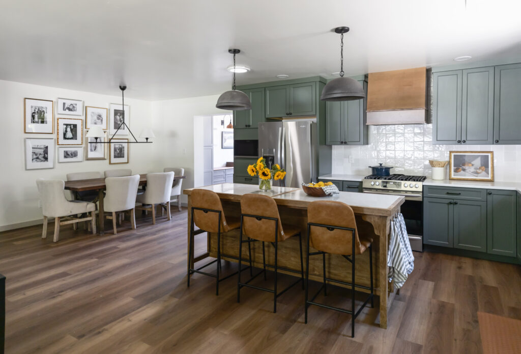 Open concept kitchen and dining room with green kitchen cabinets, leather barstools, a wood dining table with cream chairs and a gallery wall