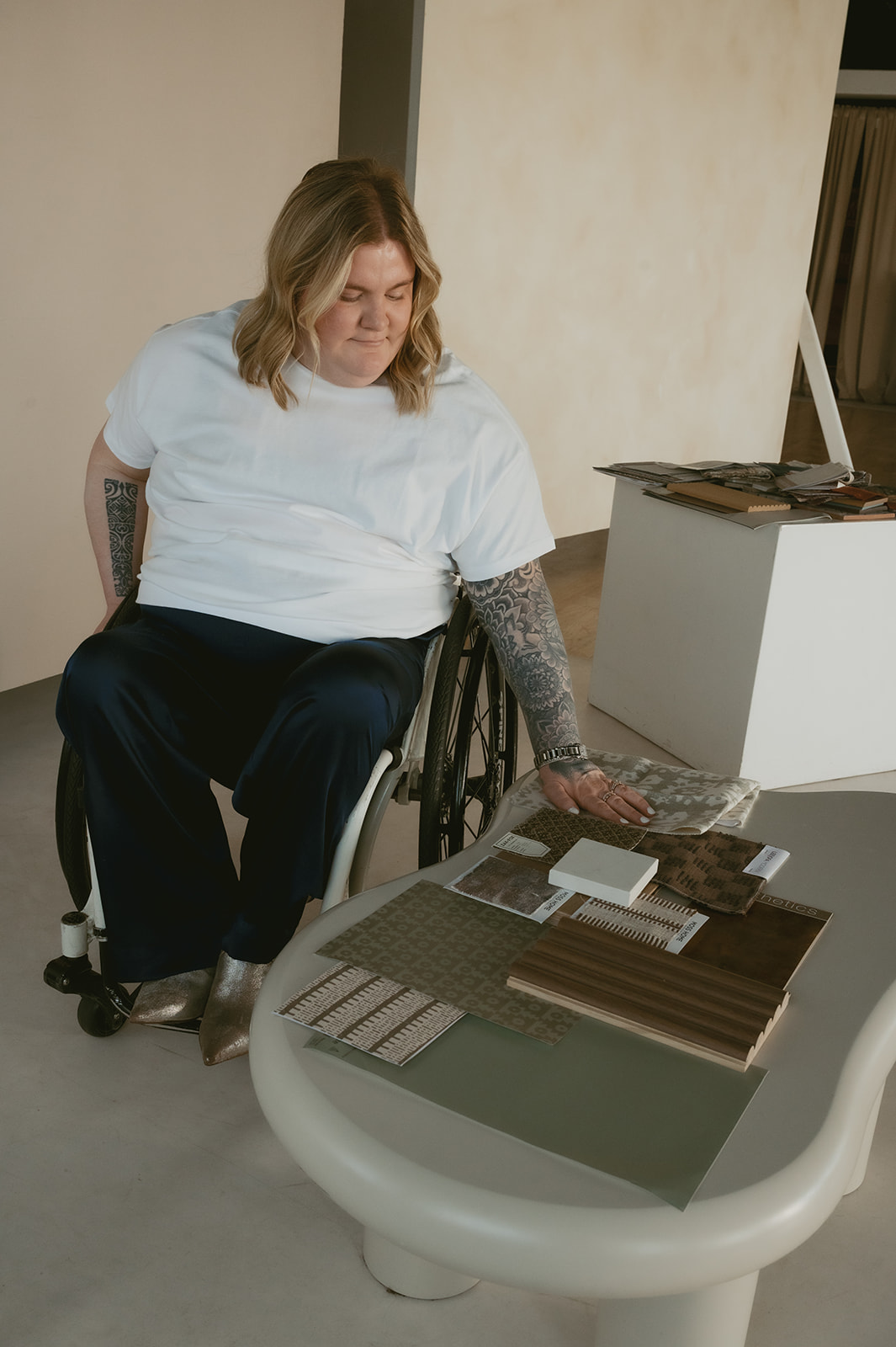 Designer with spinal cord injury in a wheelchair reviewing material samples on a table, including wood finishes, fabric swatches, and tile options.