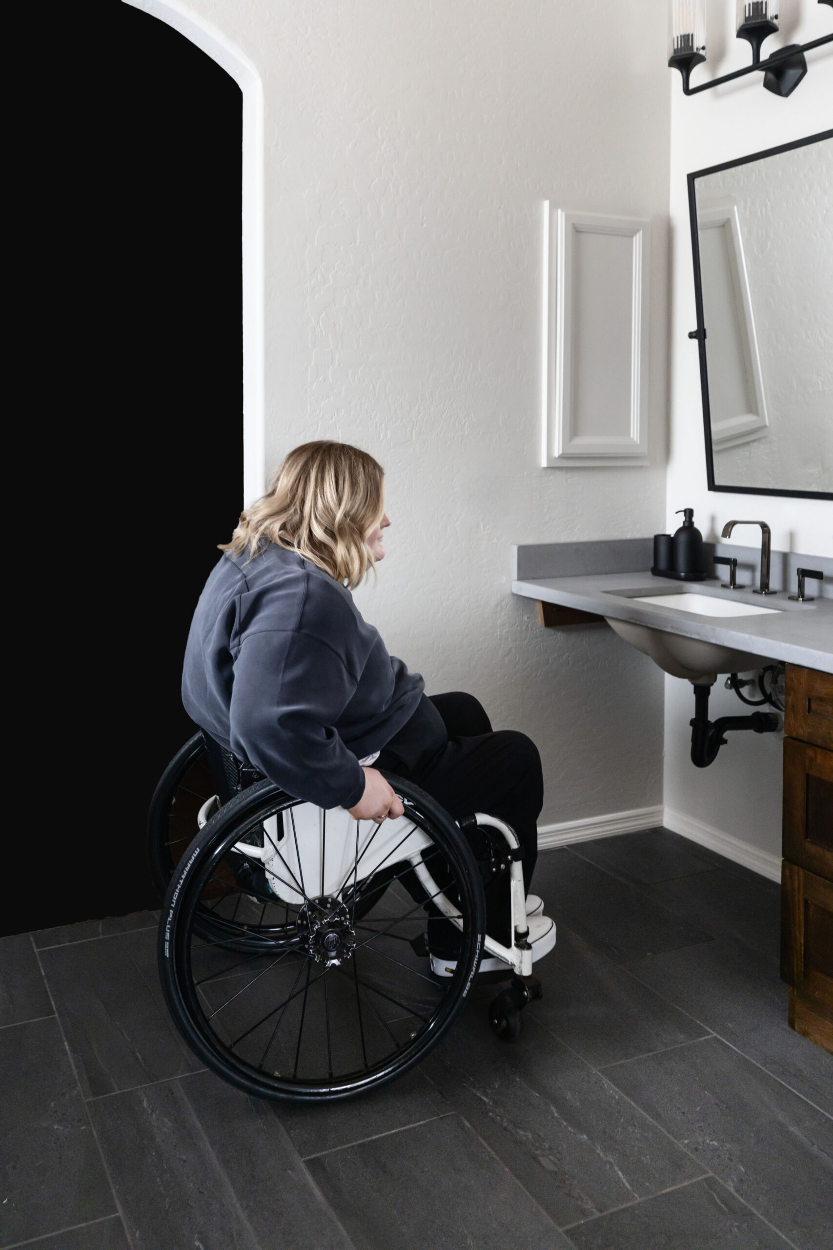 Person using a wheelchair accessing a roll-under bathroom sink, demonstrating the curb cut effect and barrier-free and accessible interior design.
