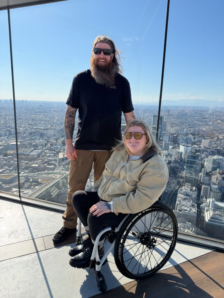 Wheelchair user enjoying an accessible observation deck, Shibuya Sky, in Japan with a companion, featuring floor to ceiling glass, level flooring, and panoramic city views that highlight inclusive travel and barrier free tourism.