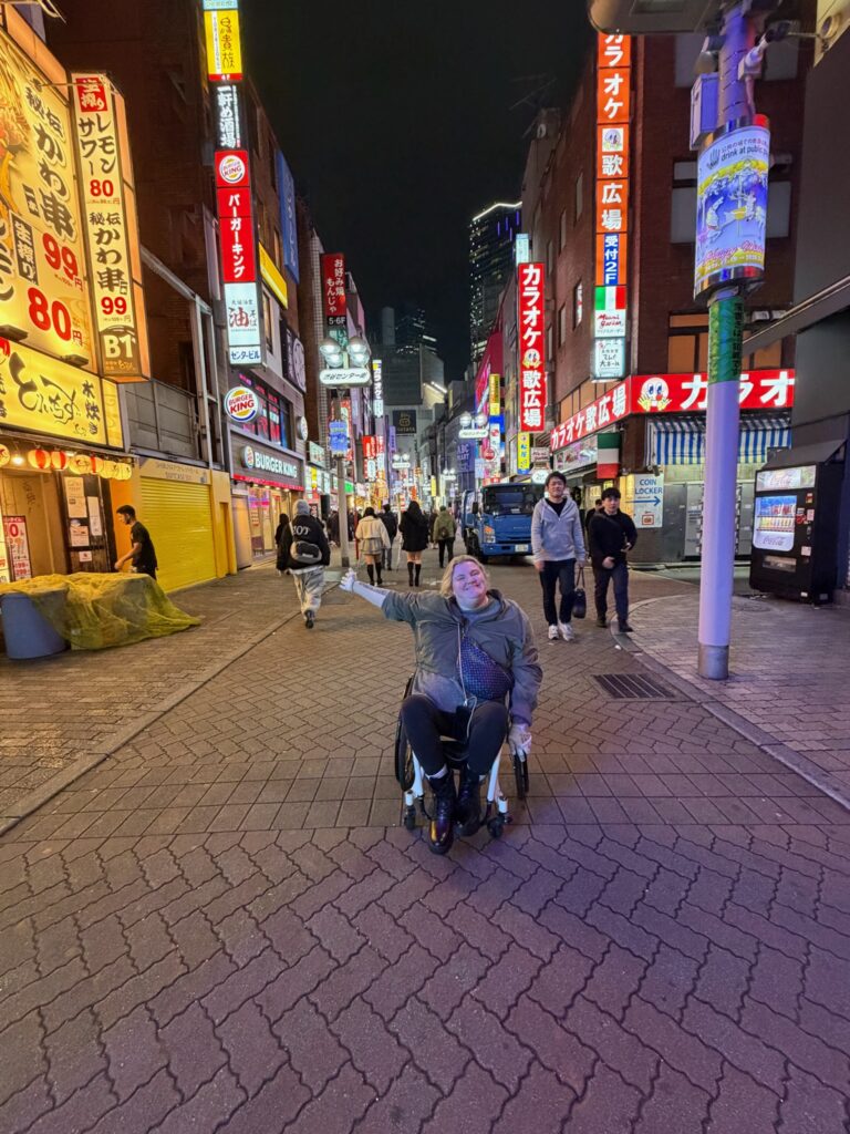 Wheelchair user enjoying accessible travel in Japan at night, navigating a vibrant Tokyo street with illuminated signage, busy pedestrian activity, and urban cityscape showcasing inclusive public spaces and mobility friendly design.