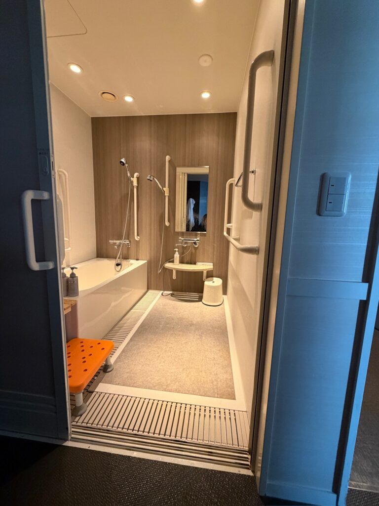 Accessible hotel bathroom in Japan with a roll in bathing area, built in tub, handheld shower, wall mounted grab bars, shower seat, wide doorway, and open floor space designed for wheelchair users and barrier free travel.