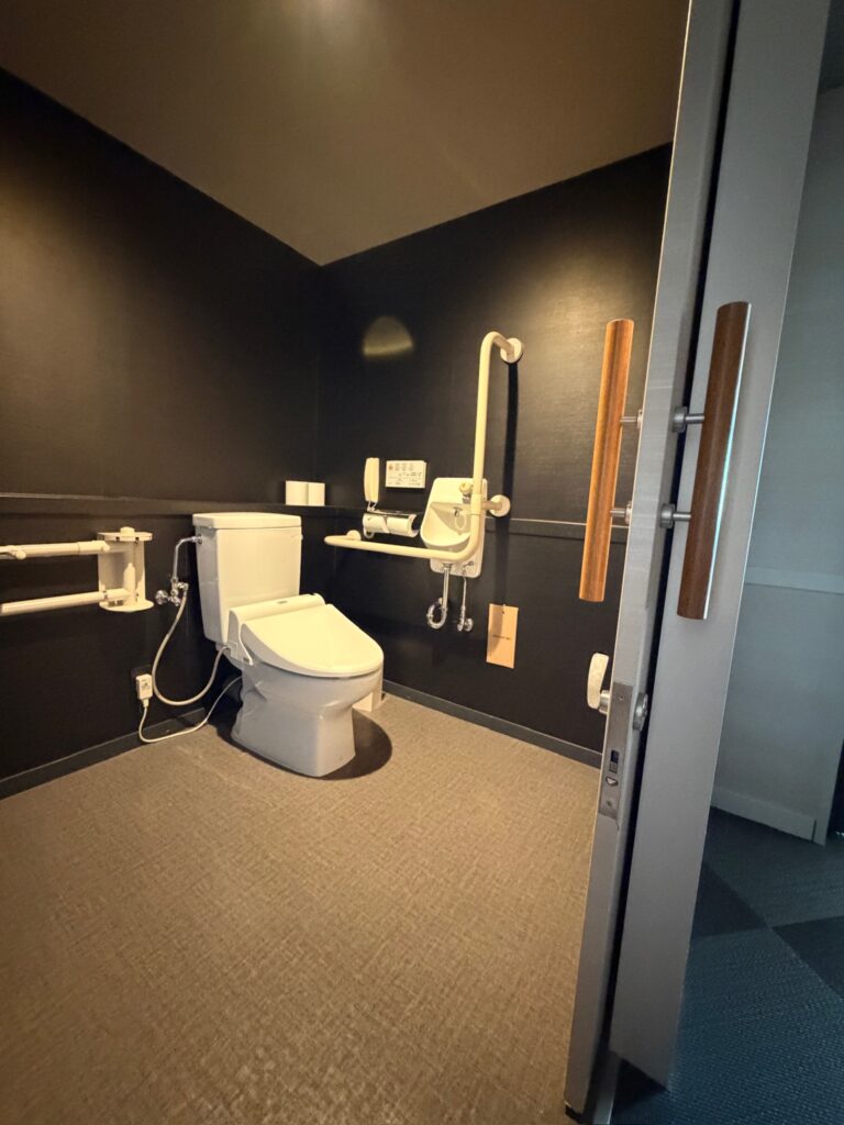 Accessible restroom in Japan featuring a bidet toilet, side and rear grab bars, transfer space, assist controls, and a wide door opening designed for wheelchair users and barrier free public bathrooms.