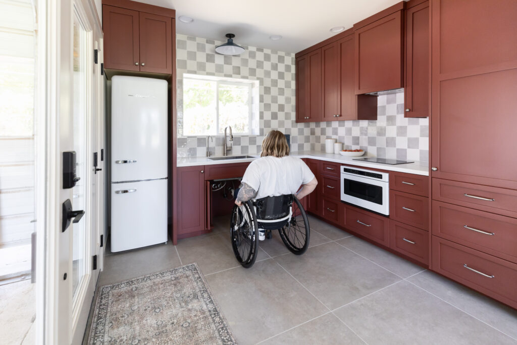 Accessible kitchen with roll under sink and cooktop, lowered work surfaces, drawer appliances, wide clearances, and a wheelchair user positioned comfortably at the counter, illustrating barrier free kitchen design that balances functionality, independence, and elevated style.